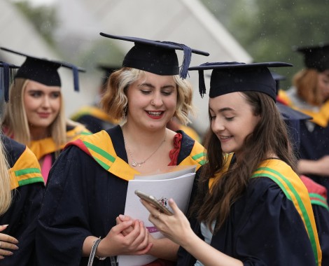 Two female students wearing cap and gowns look at a mobile phone screen.